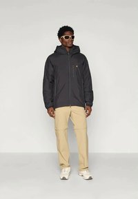 Must mustaga kapuutsiga, ees lukk ja logo tandri. Kombineeritud beežide pükstega ja valgete tossudega. Isik kannab päikeseprille.