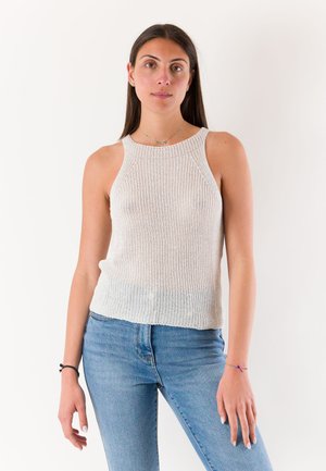 Giovane donna con capelli lunghi e scuri indossa un top bianco senza maniche in maglia e jeans blu, in piedi davanti a uno sfondo chiaro e uniforme.