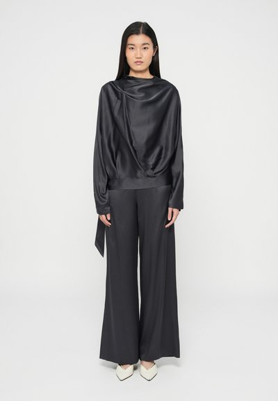 Femme debout de face portant un haut en satin noir fluide avec un col drapé, un pantalon noir à jambes larges et des chaussures blanches pointues sur fond blanc.