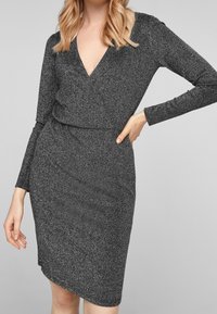 Svart långärmad klänning med djup V-ringning, tillverkad av reflekterande tyg, med en figurnära siluett och asymmetrisk nederkant.