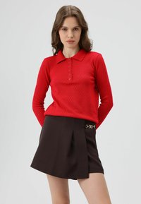 Rode polo met lange mouwen met een geperforeerd ruitpatroon en knoopsluiting. Gecombineerd met een bruine minirok met een goudkleurige kettingaccent.