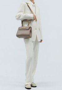 Braune Handtasche aus strukturiertem Leder mit klarer Form, Obergriff und goldfarbenen Beschlägen, gehalten neben einem cremefarbenen Anzug.