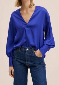 Blusa di seta blu con scollo a V e maniche lunghe a sbuffo, abbinata a jeans in denim scuro a vita alta. La blusa presenta una vestibilità rilassata e una texture liscia.