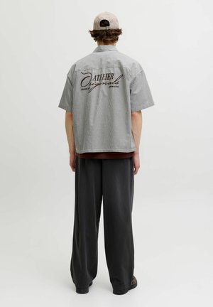 Homme portant une casquette beige, une chemise à manches courtes à carreaux avec le texte « Atelier Originals » dans le dos, un pantalon ample foncé, debout devant un fond uni.