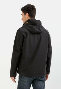 Mand iført en sort hættejakke og blå jeans, som står med ryggen til en ensfarvet lys baggrund.