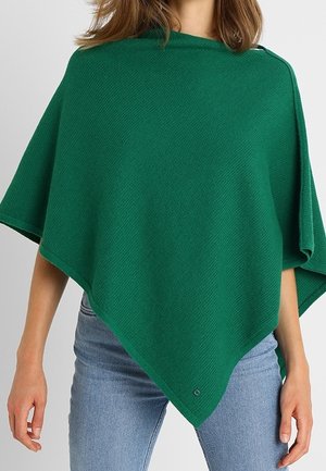 Vrouw die een groene gebreide poncho draagt over lichtblauwe spijkerbroek, staand tegen een effen lichtgrijze achtergrond.