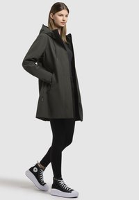 Giacca impermeabile verde scuro con cappuccio, cerniera frontale e tasche laterali. Abbinata a leggings neri e sneakers nere alte.
