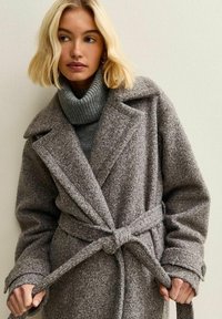 Abrigo de lana gris texturizado con amplios solapas, mangas largas y un cinturón atado en la cintura. Presenta botones en las mangas y un cuello alto con capas debajo.