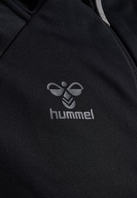 Sort atletisk zip-hættejakke med en glat overflade, der har et reflekterende Hummel-logo på venstre brystområde.