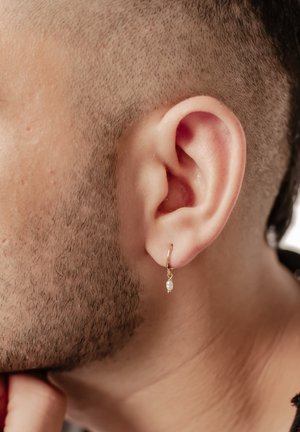 Goldene Creolen mit einem kleinen Perlenakzent, die am Ohrläppchen einer Person mit kurzem, eng geschnittenem Haar und Stoppeln befestigt sind.