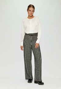 Witte blouse met lange mouwen en een gathered halslijn, gecombineerd met zwart-wit geruite wijde broek; glanzende zwarte plateau schoenen.