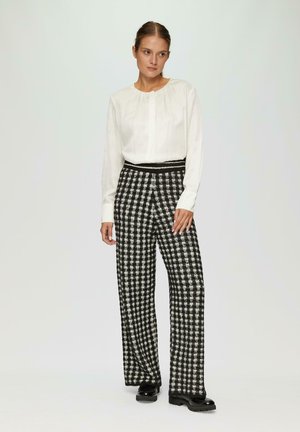 Witte blouse met lange mouwen en een gathered halslijn, gecombineerd met zwart-wit geruite wijde broek; glanzende zwarte plateau schoenen.