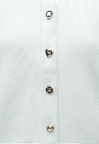 Világoskék kötött kardigán négy díszgombbal: ezüst kerek, arany szív, fekete virág és arany csengő alakú. Puha tapintású.