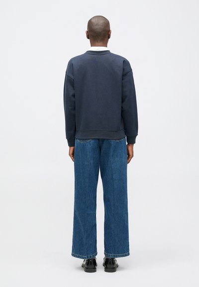 Jūras spēku džemperis ar noapaļotu apkakli un ribinātām piedurknēm, saskaņots ar platiem zila džinsa kautiem. Gluda tekstūra un brīvs piegriezums. Redzami melni apavi.