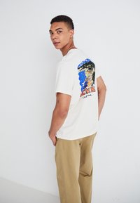 Cream cotton t-shirt with a colorful graphic design on the back featuring blue, green, and red elements, paired with light beige pants.