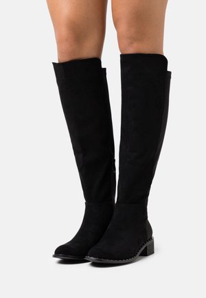 Bottes noires au-dessus du genou en daim, avec un bout arrondi, une surface texturée et des accents décoratifs cloutés le long de la semelle.