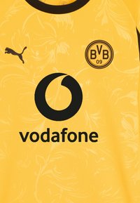 Rumena nogometna majica Borussia Dortmund z črnim logotipom Puma, grbom kluba in logotipom sponzorja Vodafone, ki vsebuje subtilen cvetlični vzorec.