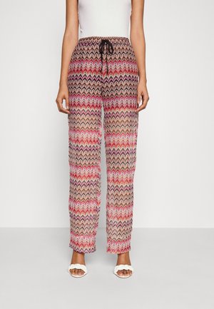 Kobieta w szerokich spodniach w wielokolorowy zygzakowaty wzór z czarnym sznurkiem, białej bezrękawnej bluzce i białych plecionych sandałach na odkryte palce.