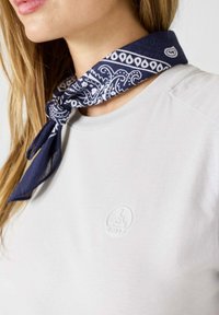 Pañoleta de estilo bandana en color azul marino con un patrón de paisley blanco, atada alrededor del cuello, usada con una camiseta de cuello redondo de tejido suave en gris claro que presenta un logo embosado.
