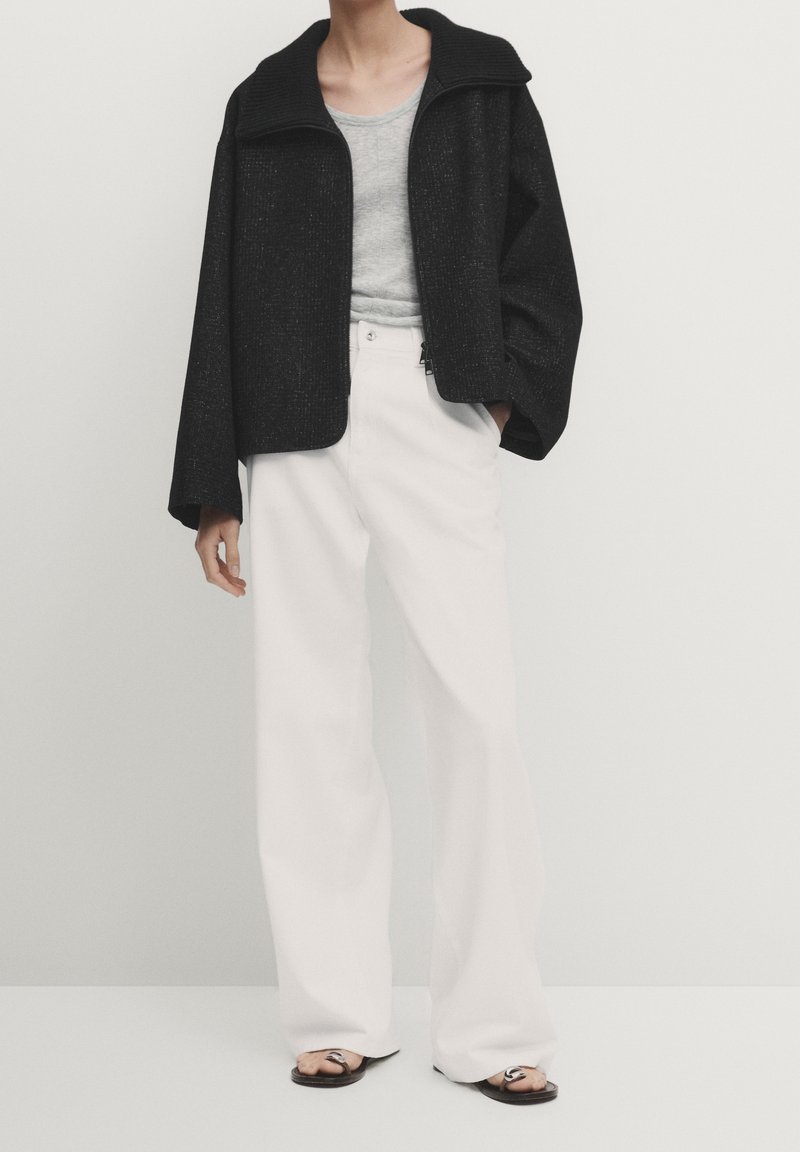 Personne portant une veste noire ample, un haut gris, un pantalon blanc large et des sandales noires, debout devant un fond clair uni.