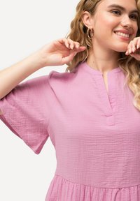 Rosa Kurzarmkleid aus strukturiertem Stoff, mit einem Schlüssellochausschnitt und einer schwingenden Silhouette, die am Taillenbereich geraffte Details aufweist.