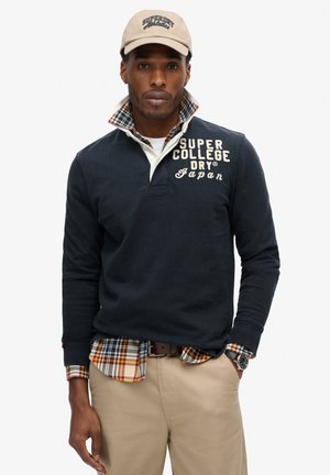 Navy long-sleeve polo with a contrasting white collar and embroidered text. Red and beige plaid shirt underneath. Casual tan trousers and watch.