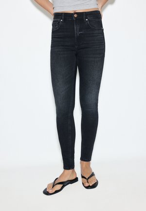 Persoon die hoge zwarte skinny jeans en zwarte slippers draagt met een lichtgrijs cropped top, staand tegen een witte achtergrond.