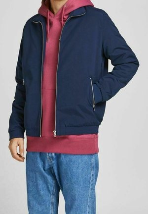 Mann trägt eine marineblaue Bomberjacke mit Reißverschluss über einem burgunderfarbenen Kapuzenpullover und blauen Jeans, steht mit einer Hand in der Tasche.