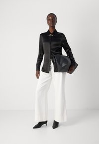 Camicia button-up in satin nero con colletto e accessorio dorato, abbinata a pantaloni bianchi larghi e décolleté neri a punta. Borsa nera in mano.