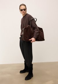 O persoană îmbrăcată cu ochelari de soare, pulover maro închis, pantaloni negri și cizme, care ține o geantă de călătorie mare din suede maro închis pe umăr.