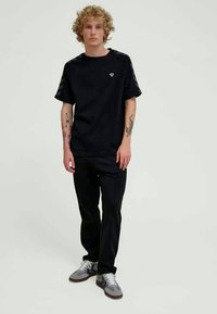 Μαύρο κοντομάνικο t-shirt με μικρό λογότυπο καρδιάς, συνδυασμένο με μαύρο παντελόνι και γκρι αθλητικά παπούτσια με μπλε λεπτομέρειες. Λεία υφή υφάσματος.