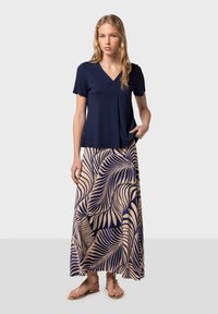 Marinblå V-ringad topp med korta ärmar, matchad med en lång kjol med beige bladmönster på en mörk bakgrund, samt platta sandaler i guld.