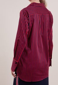 Camicia a righe rossa e blu con collo a punta, maniche lunghe e orlo leggermente curvo. Presenta un yoke sul retro.