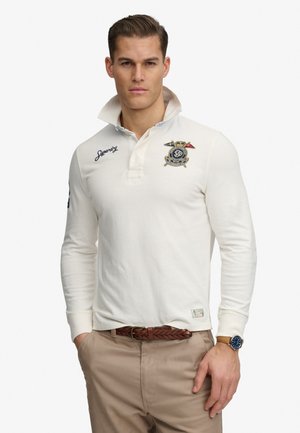 Man draagt een witte polo met lange mouwen met geborduurde logo's, beige broek, bruine gevlochten riem en horloge, staand met handen in de zakken.