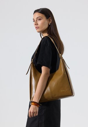 Femme aux longs cheveux bruns portant une robe noire, un grand sac à épaule en cuir marron et un bracelet épais en ambre.