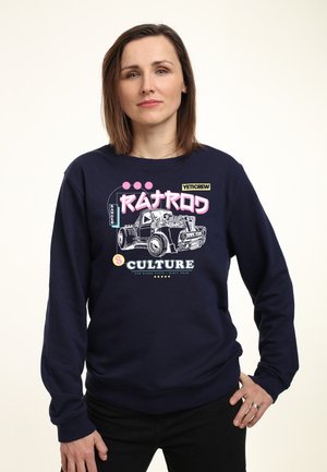 Kobieta z prostymi brązowymi włosami w granatowej bluzie z nadrukiem ciężarówki i napisem „RAMATROD CULTURE” różowymi i białymi literami.
