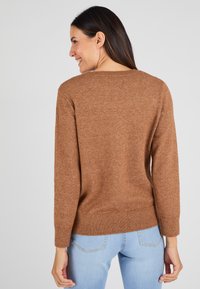 Maglione marrone, texturizzato, con scollo rotondo, maniche lunghe e orlo a coste, abbinato a jeans blu chiaro. Foto scattata da dietro.
