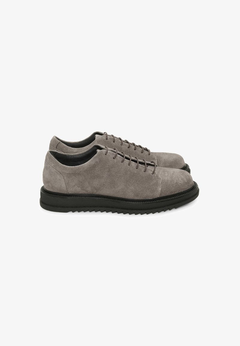 Paire de chaussures à lacets en daim gris avec semelles en caoutchouc noires à motif zigzag, vues de côté sur un fond blanc.