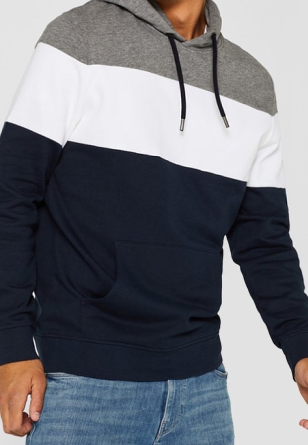 Homme portant un sweat à capuche color-blocké avec des sections gris, blanc et bleu marine, associé à un jean bleu clair, debout devant un fond uni.