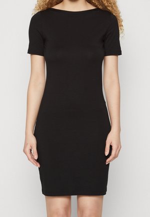 Kobieta w obcisłej, czarnej sukience z krótkim rękawem stoi na tle jednolitego jasnego tła.