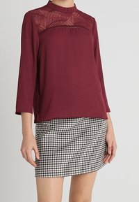 Bordeauxblus med spetsdetaljer vid oket, trekvartslånga ärmar; ihop med en svartvit rutig minikjol. Slät textur.