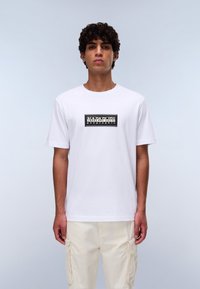 Hvid bomulds t-shirt med korte ærmer, med et sort logo print centreret på brystet. Pasformen er afslappet, og teksturen er glat.