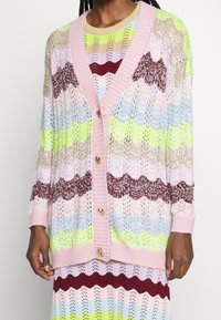 Monivärinen neulottu villatakki, jossa on aaltoileva kuvio, pinkki ribbineulekaulus ja sydämenmuotoiset napit, päällä yhteensopivan mekon kanssa.