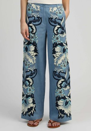Pantalon bleu à jambe large avec de grands motifs floraux en noir, blanc et turquoise, porté avec des sandales marron et un haut blanc.