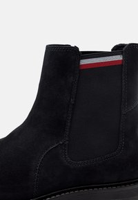 Bottine en daim noir avec panneau latéral élastique orné de rayures rouges et blanches, surpiqûres détaillées et semelle basse.