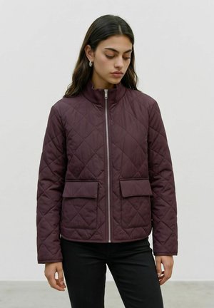 Jeune femme portant une veste matelassée violet foncé avec fermeture éclair à l'avant et deux poches à rabat, debout devant un fond clair uni.