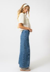 Un haut court texturé de couleur claire avec des manches courtes, associé à une longue jupe en denim bleue taille haute. La vue de côté montre des baskets décontractées.