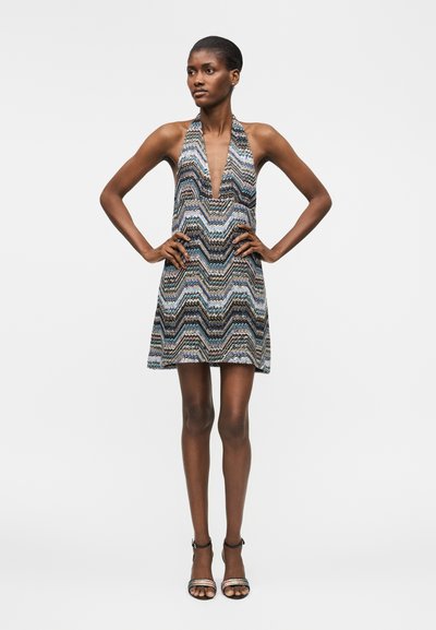 Λεπτή γυναίκα με κοντά μαλλιά που φοράει μίνι φόρεμα χωρίς μανίκια με V-λαιμόκοψη και ζιγκ-ζαγκ μπλε, μαύρο και άσπρο σχέδιο, στέκεται με τα χέρια στη μέση.