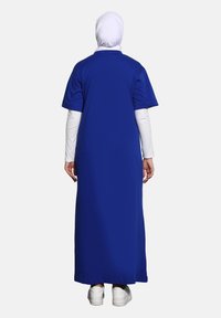 Dlouhá modrá šaty s krátkými rukávy, hladká tkanina a rovný střih, nošené přes bílé tričko s dlouhými rukávy. Bílé tenisky dokončují vzhled.