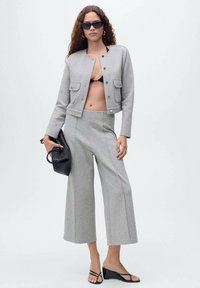 Veste grise courte avec poches avant, associée à un pantalon large gris. Le modèle porte un sac à main noir texturé et des lunettes de soleil noires.